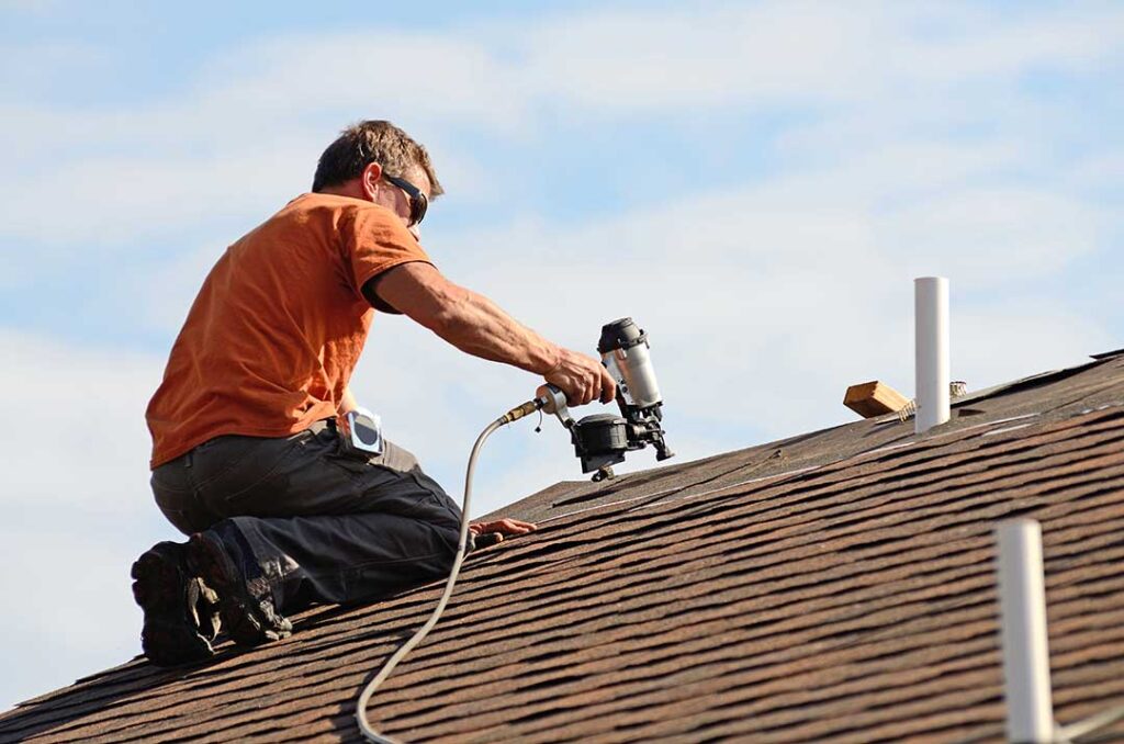 A person inspecting a pitched shingle roof for damage after a strong storm with scattered debris visible.